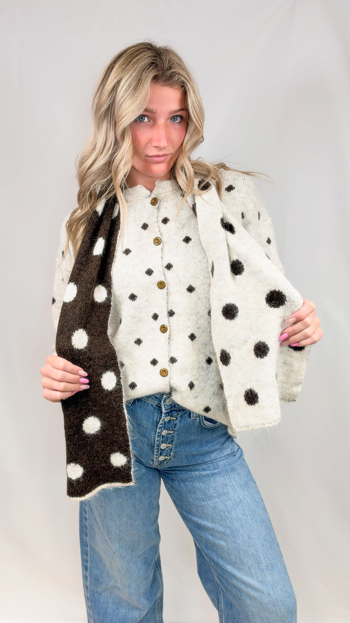 Woman wearing oatmeal color cardigan with brown polka dots and matching scarf with jeans on plain background