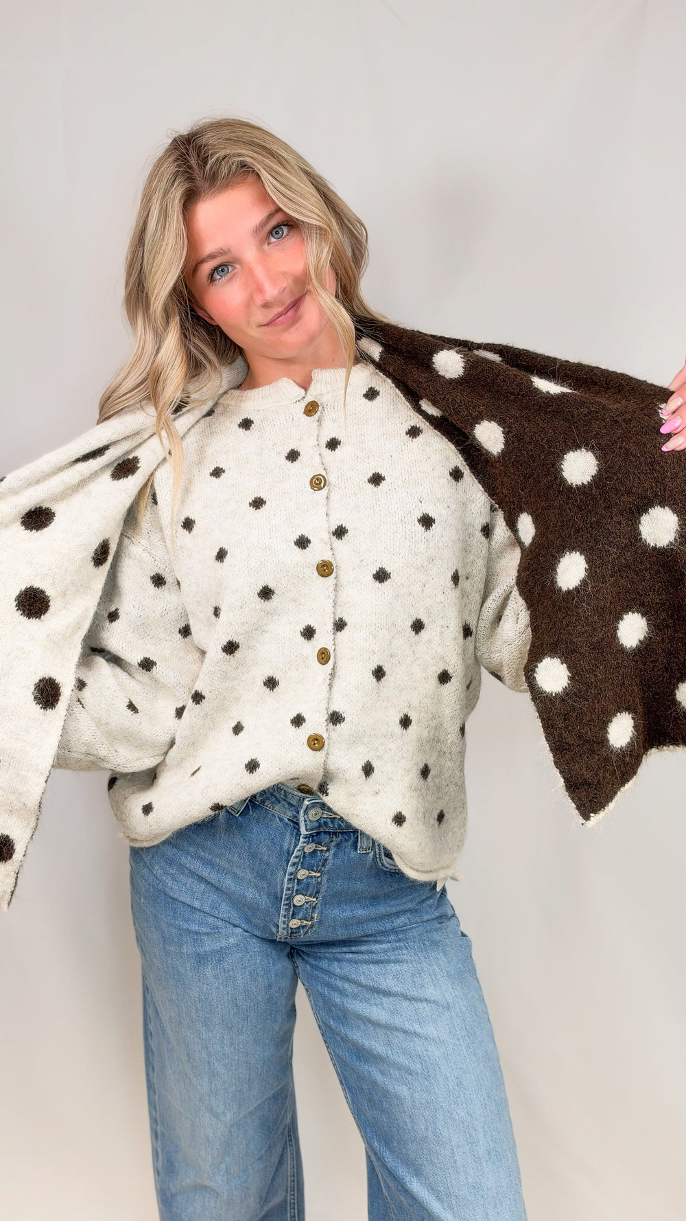 Woman wearing oatmeal cardigan with brown polka dots and matching scarf with jeans on plain background