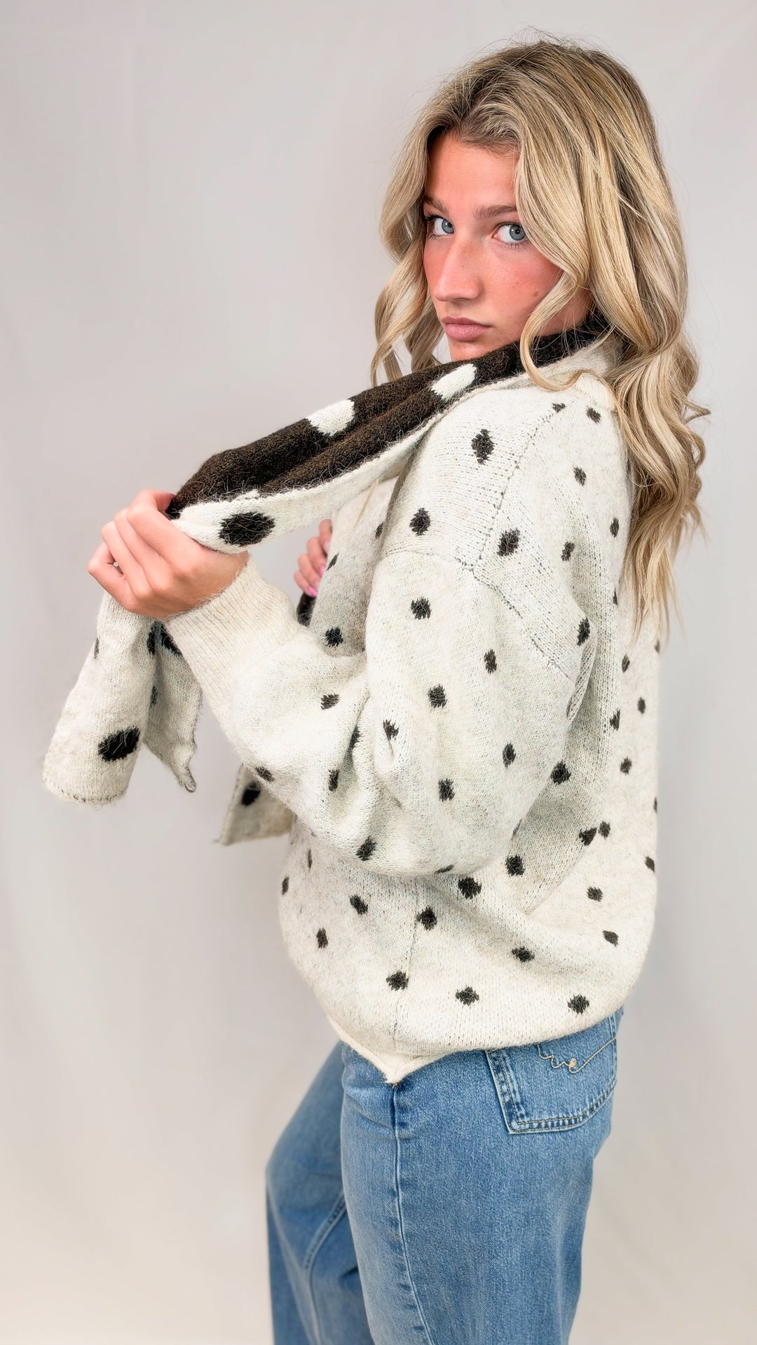 Woman wearing oatmeal cardigan with brown polka dots and matching scarf with jeans on plain background
