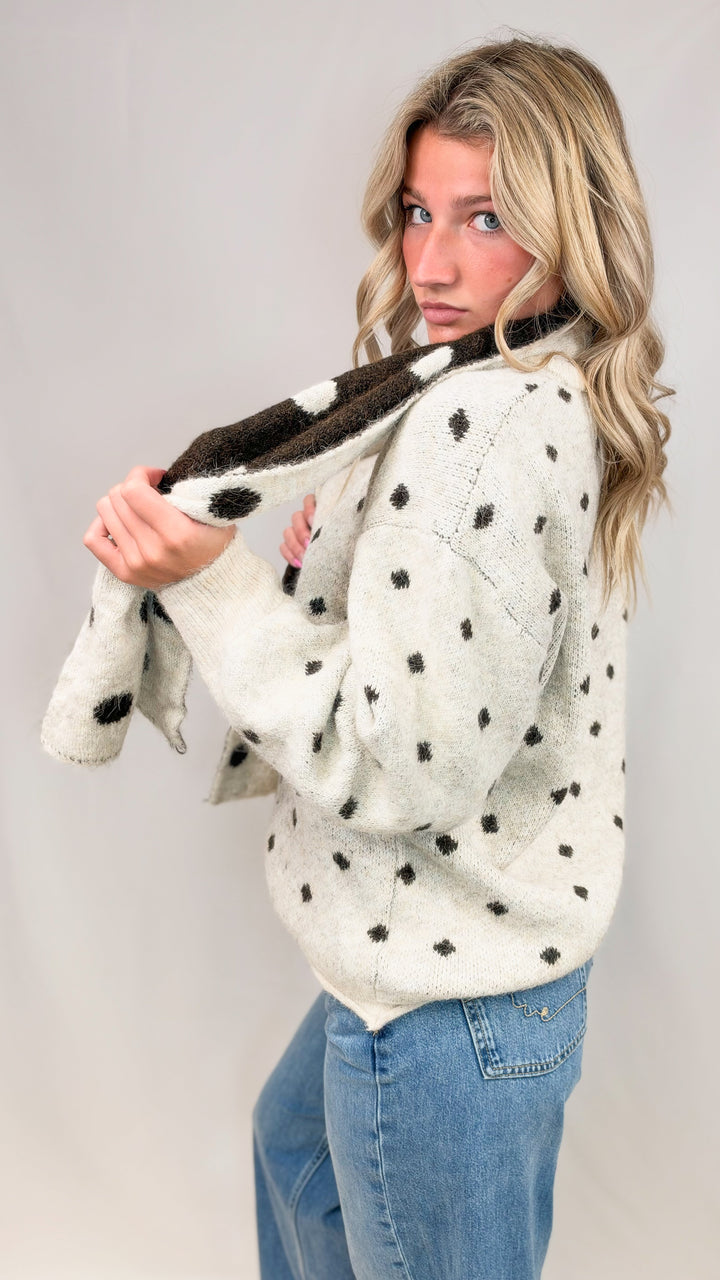 Woman wearing oatmeal cardigan with brown polka dots and matching scarf with jeans on plain background