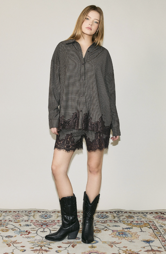 A woman stands indoors wearing an oversized black-and-white patterned shirt with matching shorts featuring a lace hem, paired with black cowboy boots, against a plain light wall and patterned rug.