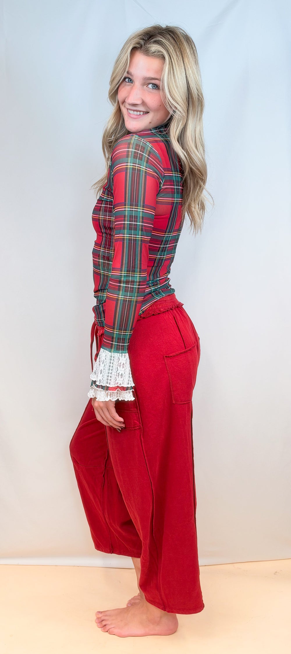 Woman shown from side view wearing red tartan sheer top with red pants on plain background