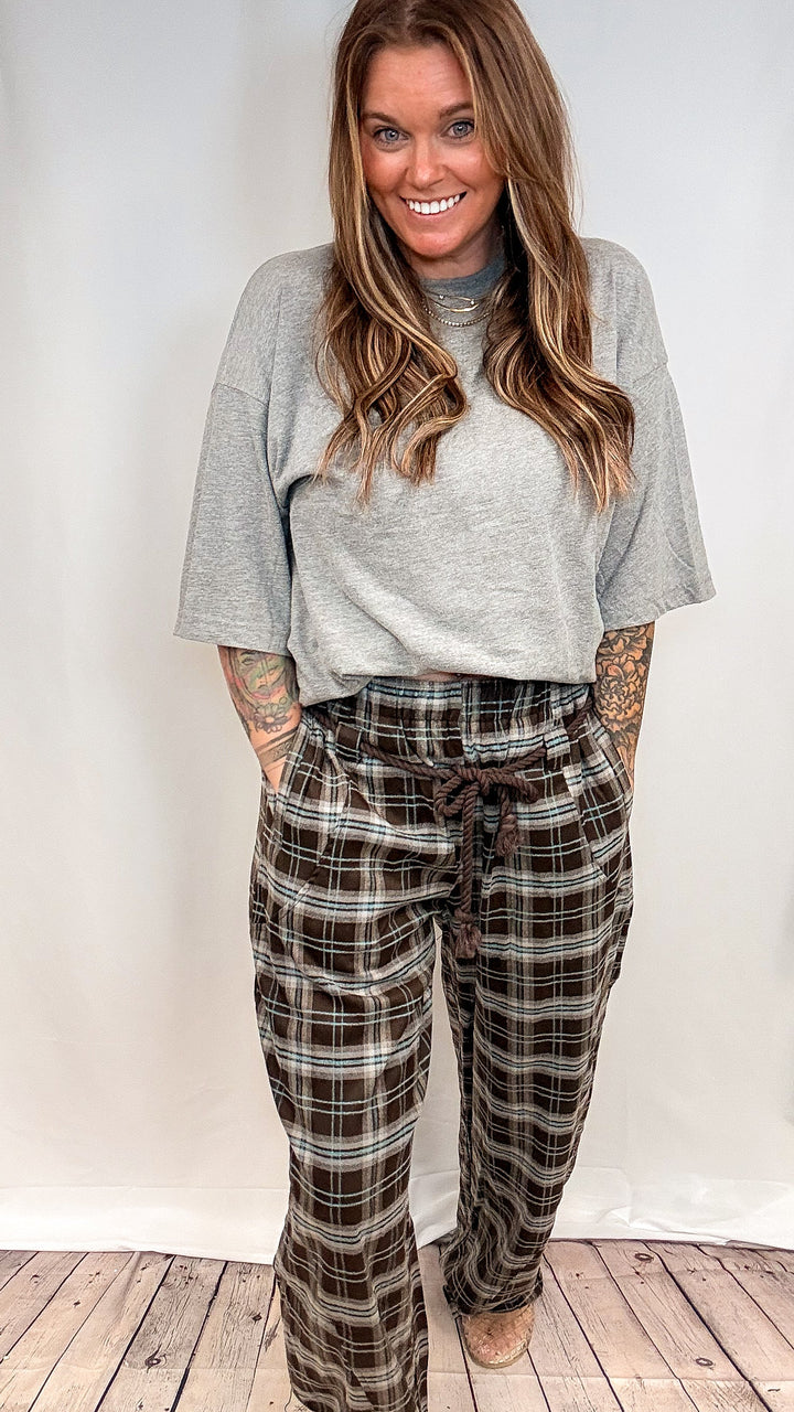 A woman modeling a light gray crewneck sweatshirt and brown plaid drawstring pajama pants against a white studio backdrop with a wooden floor.