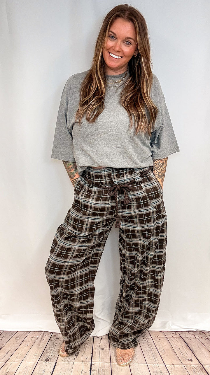 A woman modeling a gray short-sleeve t-shirt and brown plaid wide-leg pants against a white studio backdrop with a light wood floor.
