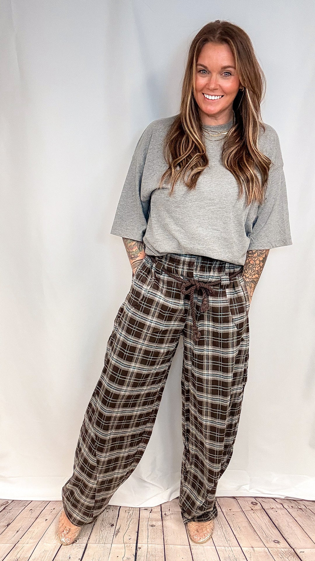 A woman modeling a pair of brown and beige plaid wide-leg lounge pants with a drawstring waist, paired with a gray t-shirt, standing against a white studio backdrop with a light wood floor.