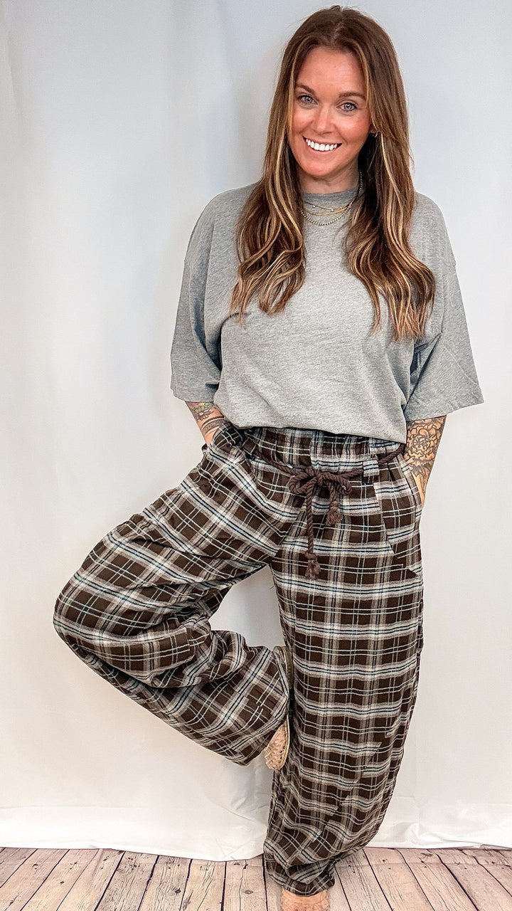 A woman modeling a pair of brown and beige plaid wide-leg pants with a drawstring waist, paired with a gray t-shirt, standing against a white studio backdrop with a wooden floor.