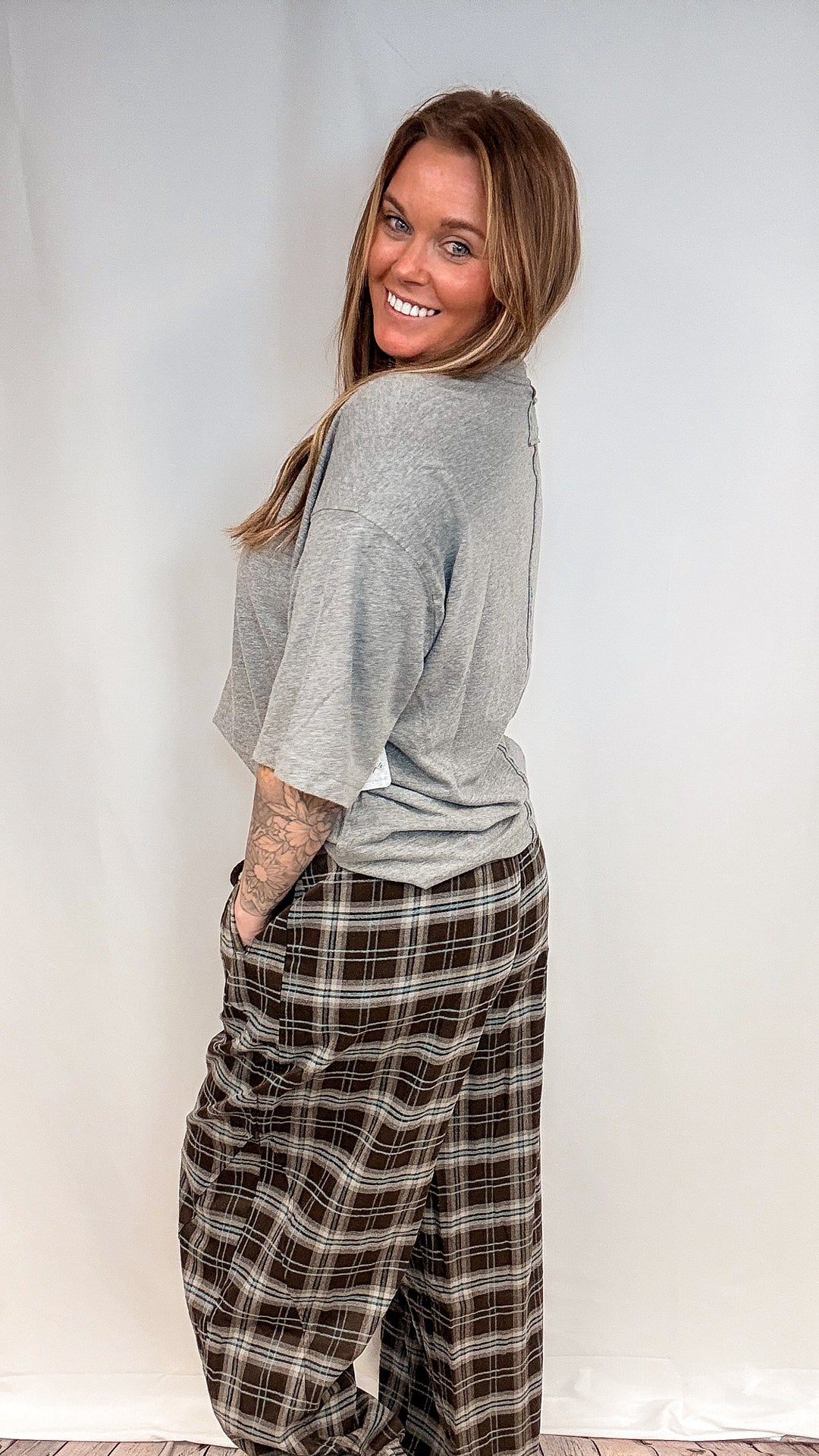 A woman wearing a light gray short-sleeve t-shirt and brown plaid pants, posing in front of a white studio backdrop.