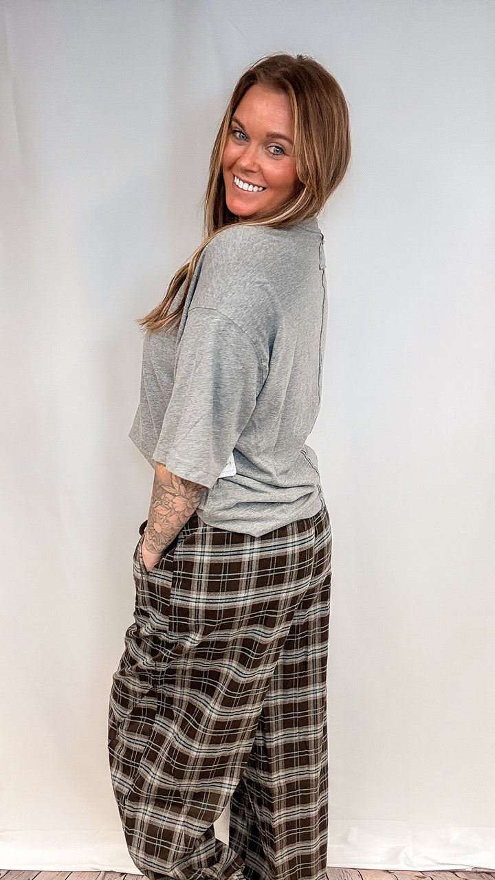 A woman wearing a light gray short-sleeve t-shirt and brown plaid pants, posing in front of a white studio backdrop.