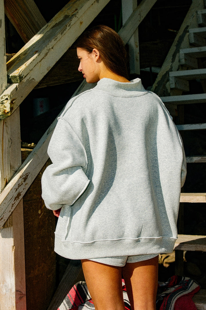 Person wearing a light gray oversized sweatshirt and shorts standing on wooden steps.
