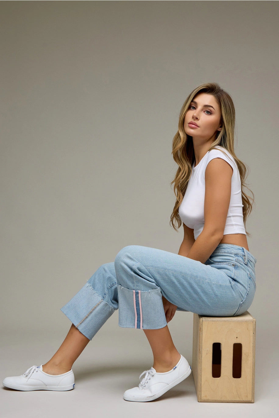Woman sitting on a wooden block wearing a white top and light blue jeans against a plain background