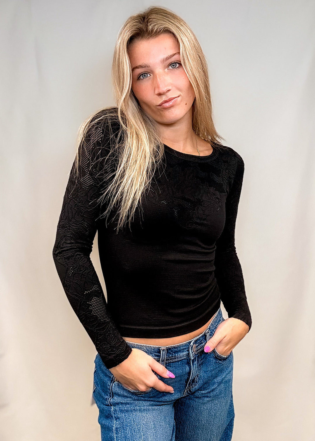 A blonde woman wearing a black long-sleeve lace top and blue jeans, standing against a light gray fabric backdrop.
