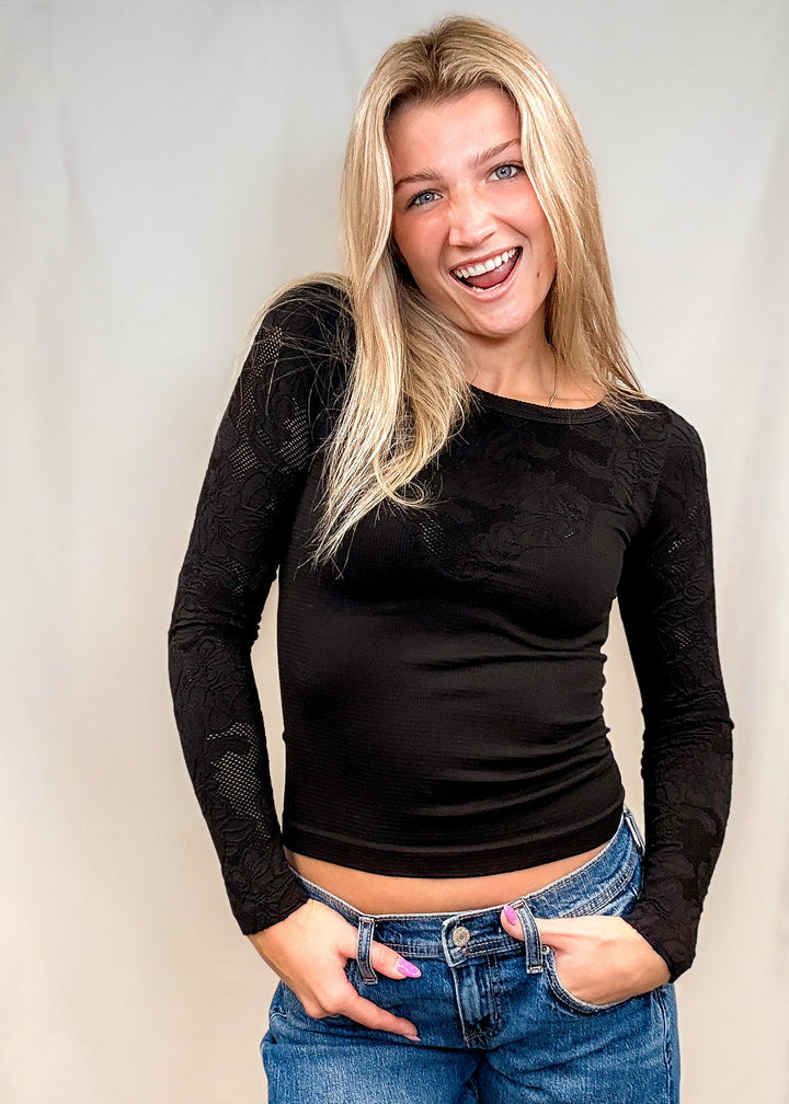 A smiling blonde woman wearing a fitted black long-sleeve top and blue jeans, standing against a plain light backdrop.