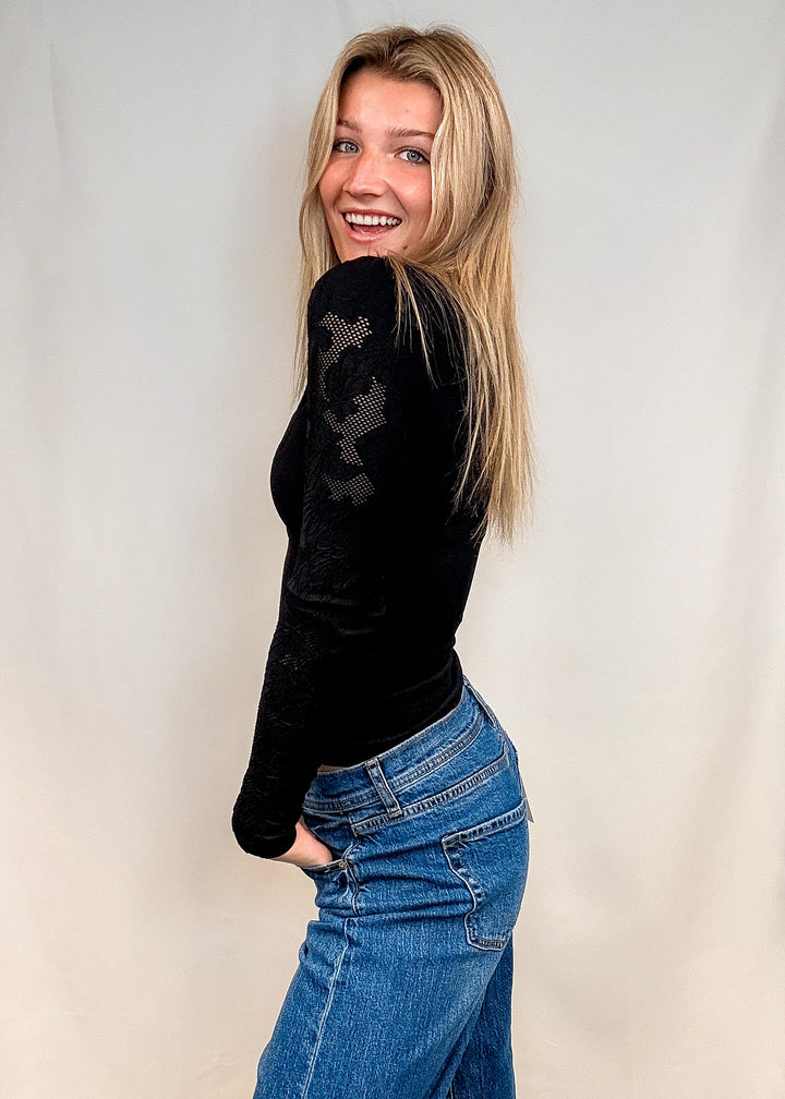 A woman with long blonde hair smiling and looking back over her shoulder, wearing a black long-sleeve knit top with sheer floral lace detailing on the back and high-waisted blue jeans, standing against a white fabric backdrop.