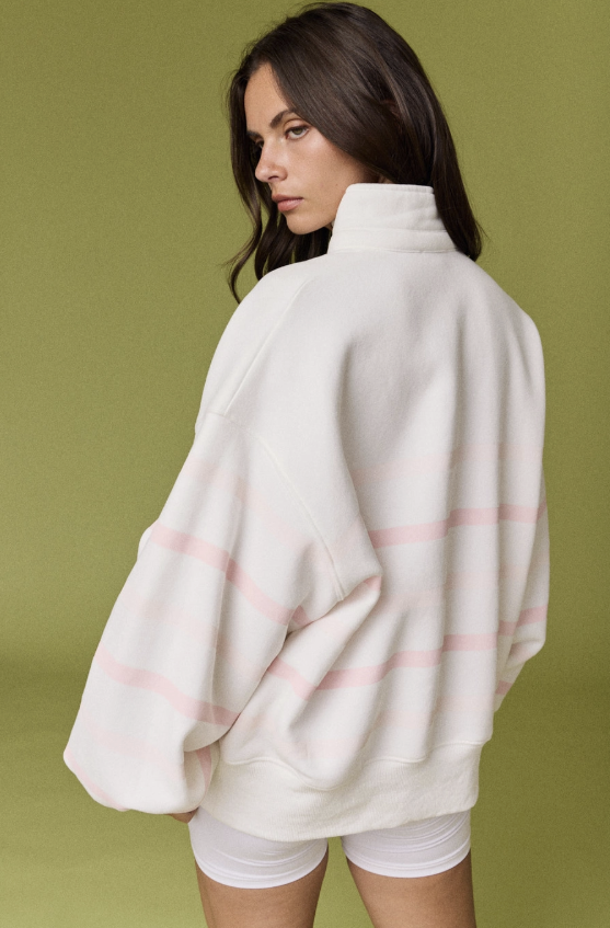 A woman wearing an oversized cream-colored fleece pullover sweatshirt with thin pink horizontal stripes, shown from a back-three-quarter angle against a solid olive green studio background.