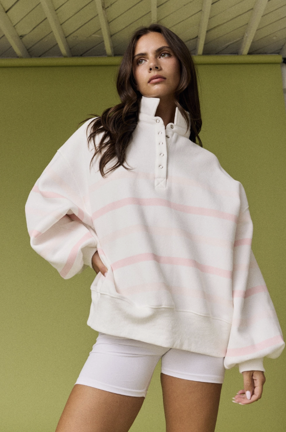 A woman wearing an oversized white sweatshirt with light pink horizontal stripes and a high button placket, paired with white fitted shorts, posed against a solid olive green wall.