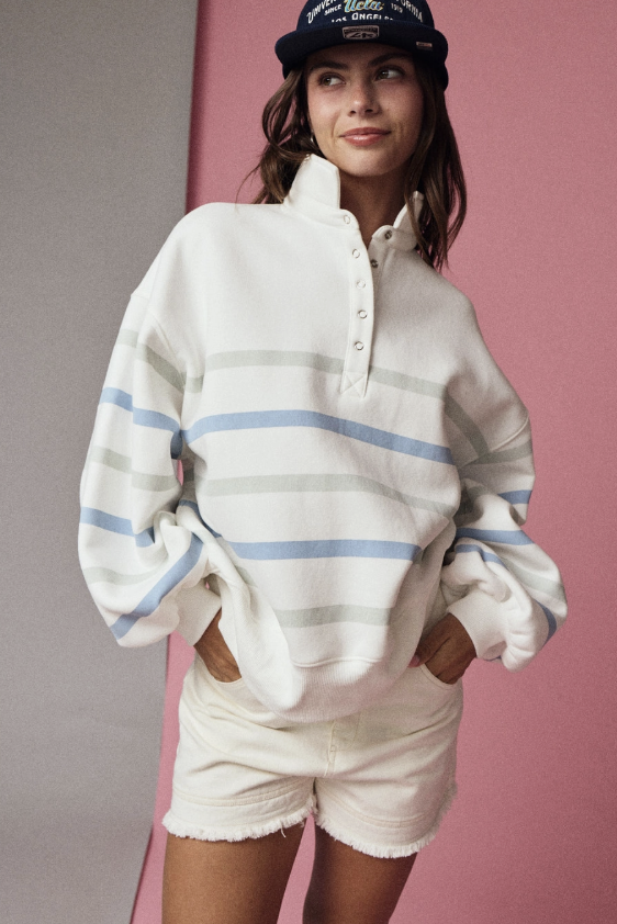 A woman wearing a cream-colored, oversized quarter-zip sweatshirt with light blue and gray horizontal stripes, paired with matching cream shorts and a black baseball cap, standing against a split pink and gray background.
