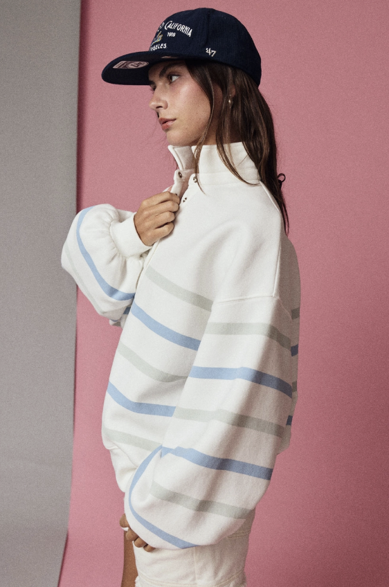A woman wearing a white quarter-zip sweatshirt with light blue and gray horizontal stripes, paired with a navy baseball cap, posed against a pink and gray studio backdrop.