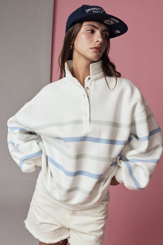 A woman wearing an oversized white half-placket pullover sweatshirt with light blue and gray horizontal stripes, paired with white shorts and a navy blue baseball cap, set against a split gray and pink background.