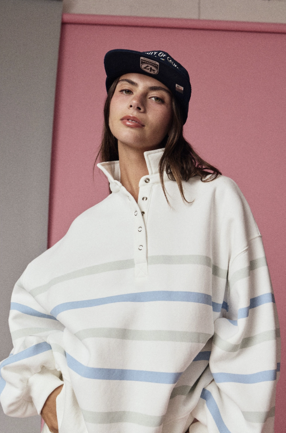 A woman wearing an oversized white long-sleeve polo shirt with light blue and gray horizontal stripes, paired with a black baseball cap, posed against a pink wall.