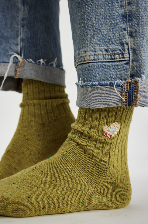 Close-up of a person wearing olive green ribbed knit socks with tiny multicolor speckles and a small embroidered logo near the cuff, paired with cuffed blue jeans on a light gray background.
