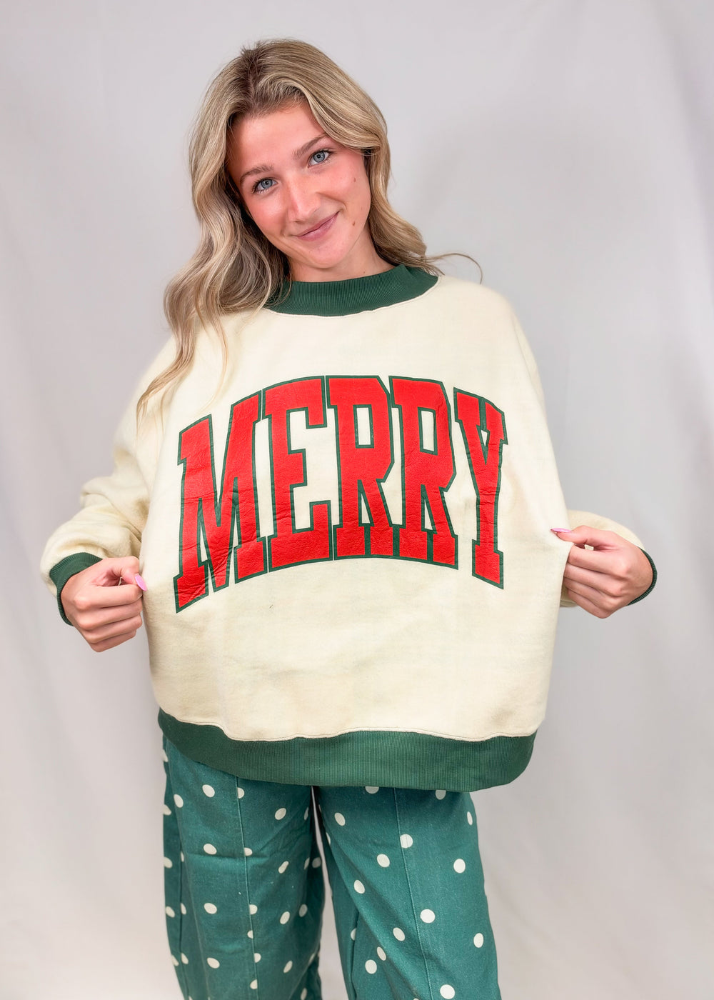 Woman wearing beige mock neck sweatshirt with 'MERRY' printed on front with green polka dot pants on plain background
