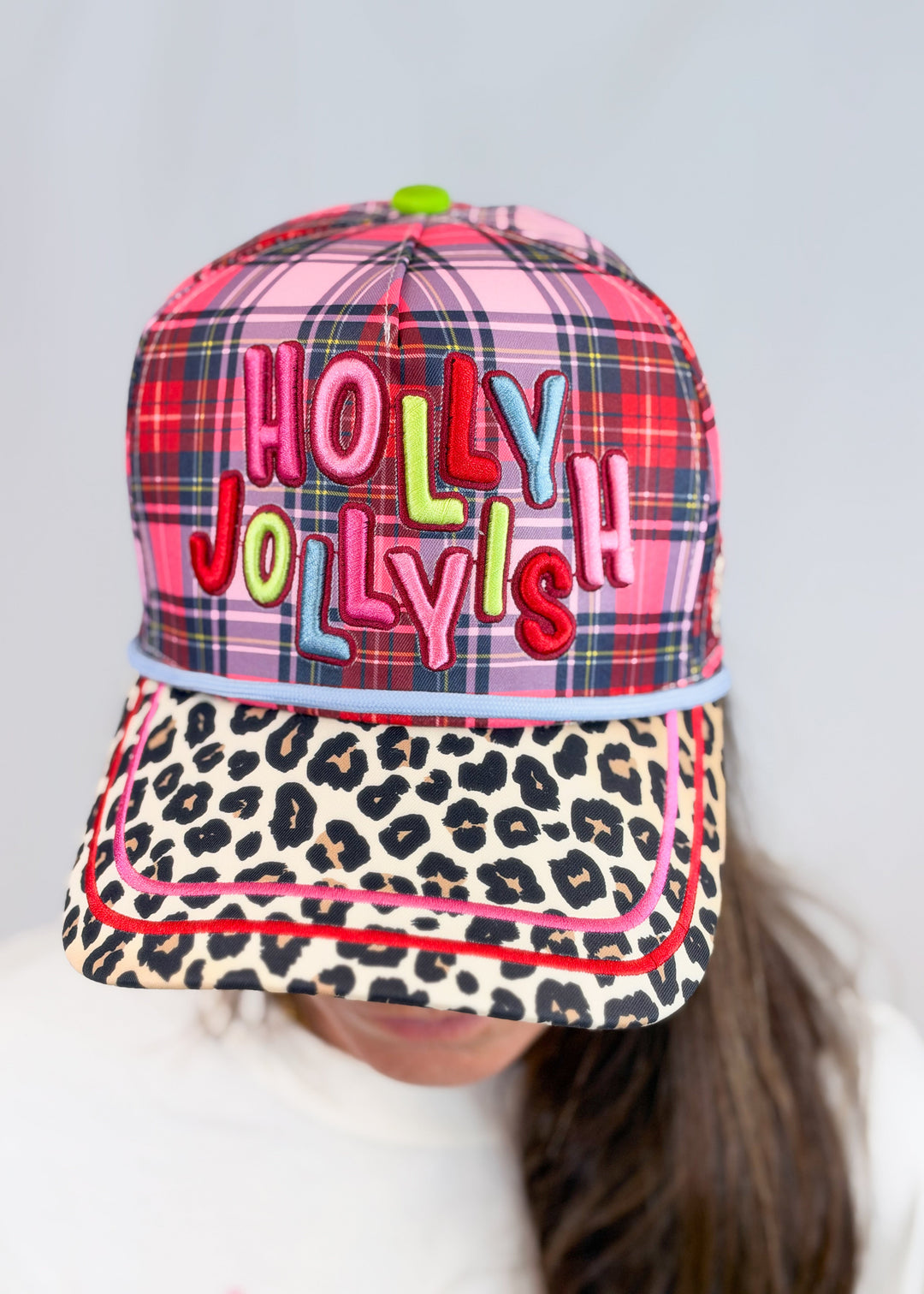 Woman wearing pink, purple and red plaid hat with 'HOLLY JOLLYISH' embroidered on front with leopard print bill on plain background