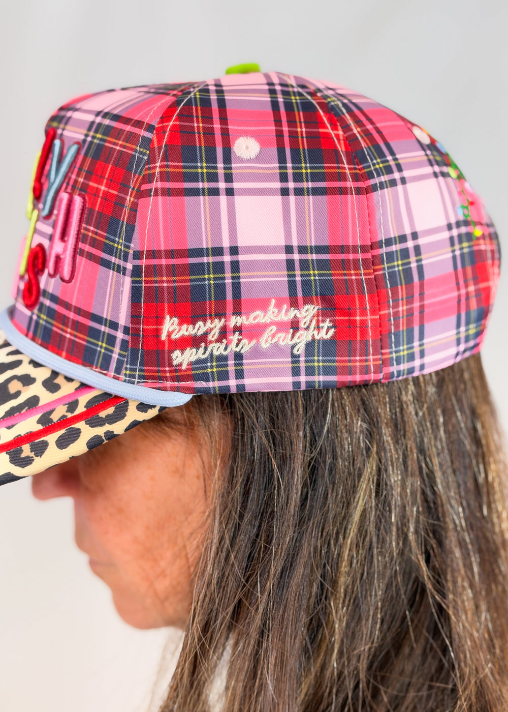 Woman wearing pink, purple, and red plaid trucker hat with 'bust making spirits bright' embroidered on side on plain background