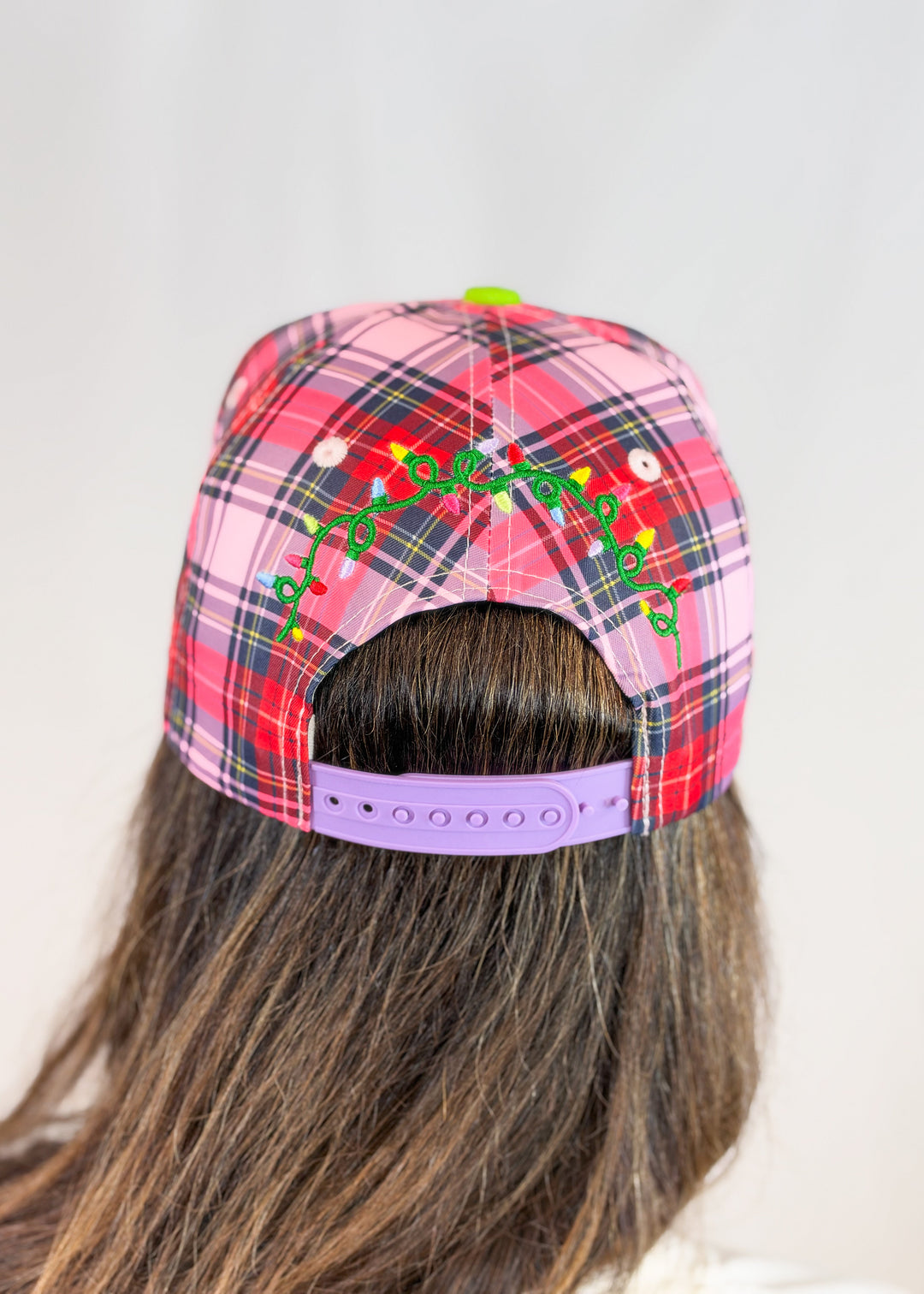 Woman shown from back wearing pink, purple, and red plaid trucker hat with christmas lights embroidered on back and purple snap on plain background