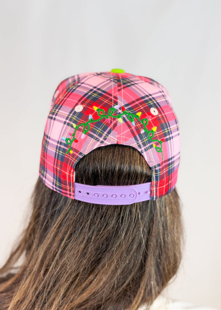 Woman shown from back wearing pink, purple, and red plaid trucker hat with christmas lights embroidered on back and purple snap on plain background