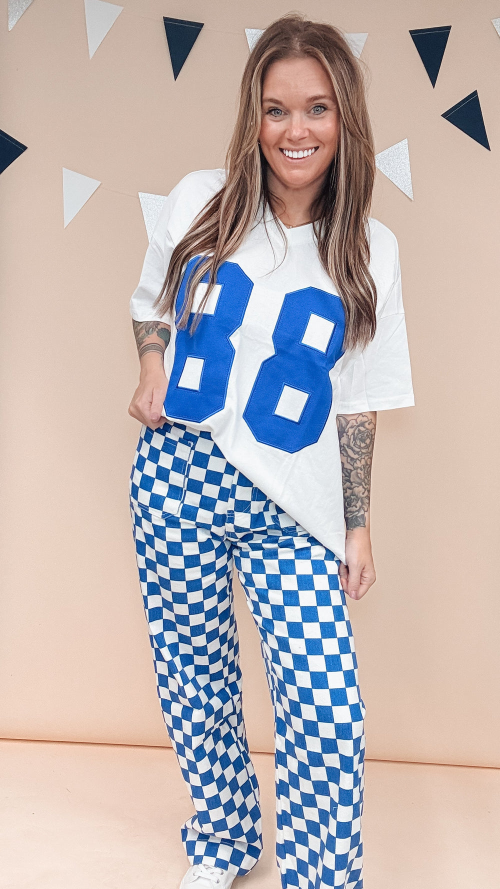 A woman modeling a white t-shirt with large blue '88' lettering and blue-and-white checkered pants, standing against a beige wall with triangular bunting.