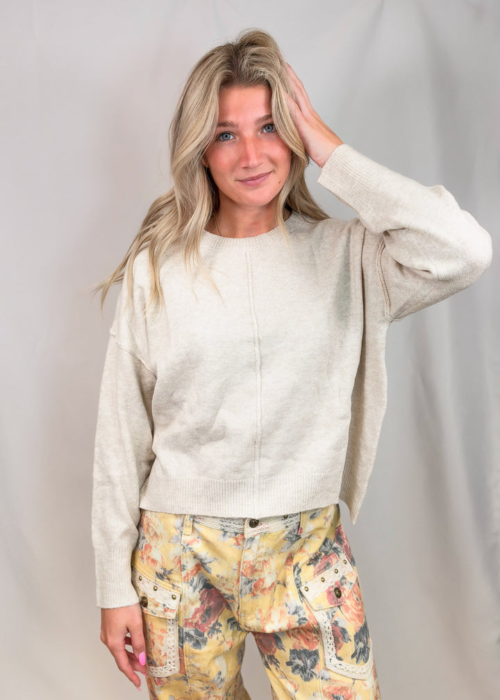 A woman modeling a cream-colored knit crewneck sweater paired with yellow floral cargo pants, standing against a light gray studio backdrop.