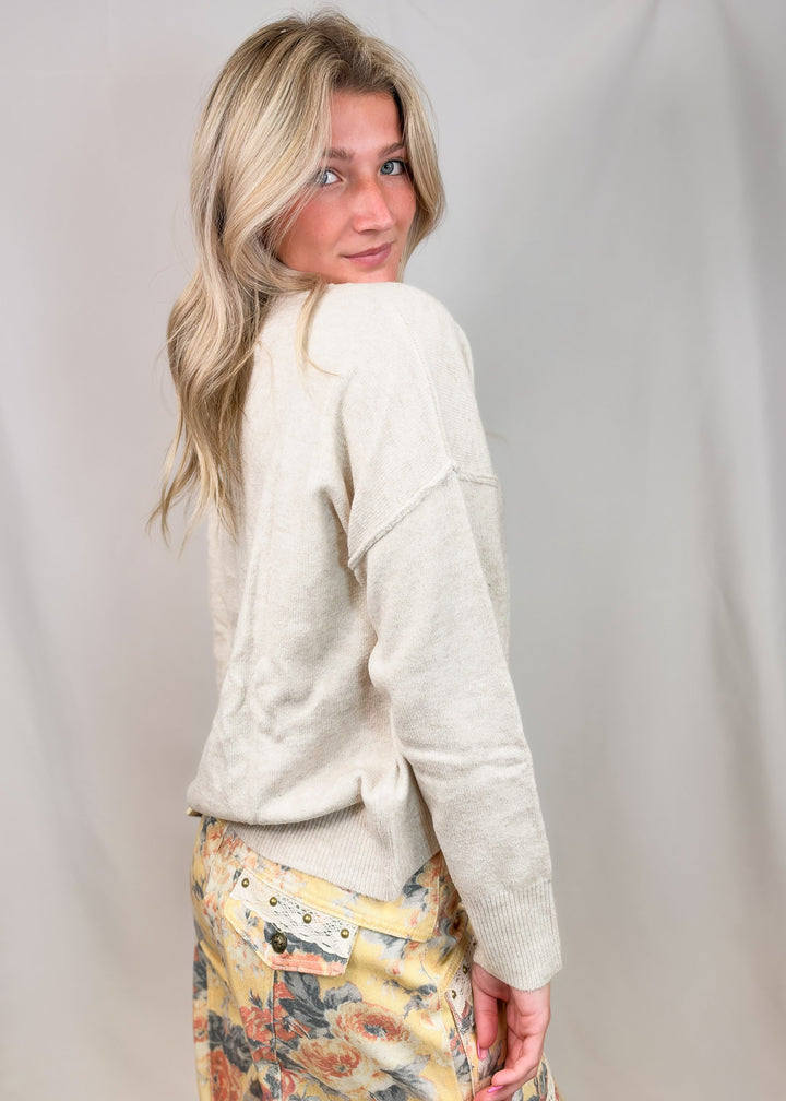 A woman with blonde hair is shown from the back wearing a cream-colored knit sweater and a yellow floral skirt, standing against a light gray fabric backdrop.