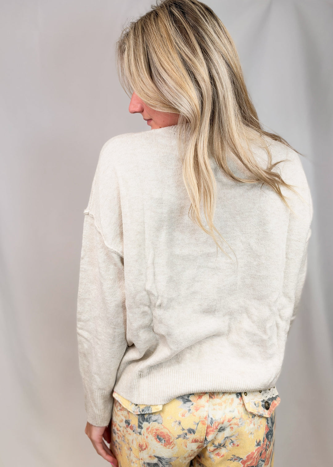 A woman shown from the back wearing a cream-colored knit sweater and yellow floral pants against a light gray studio backdrop.