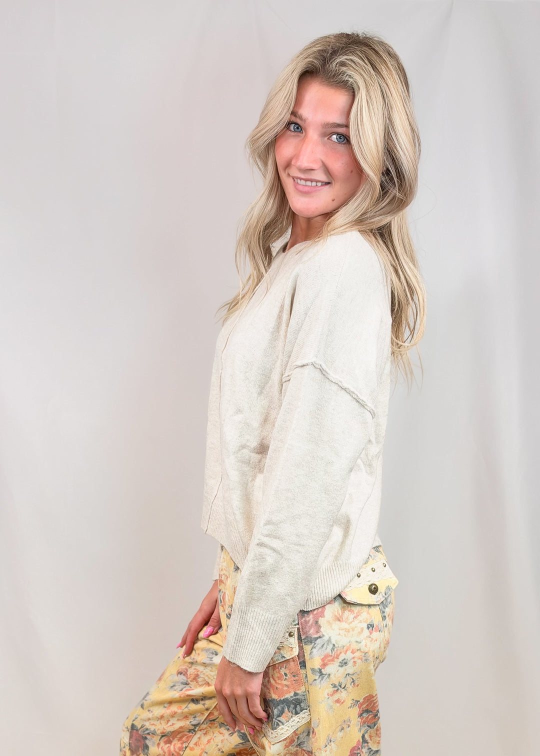 A woman modeling loose, harem-style pants with a yellow and multicolor floral print, paired with a cream-colored long-sleeve top, standing against a light beige studio backdrop.