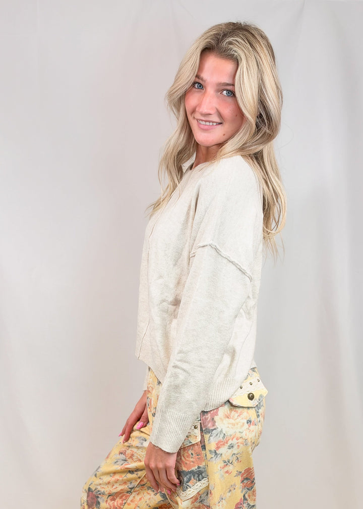 A woman modeling loose, harem-style pants with a yellow and multicolor floral print, paired with a cream-colored long-sleeve top, standing against a light beige studio backdrop.