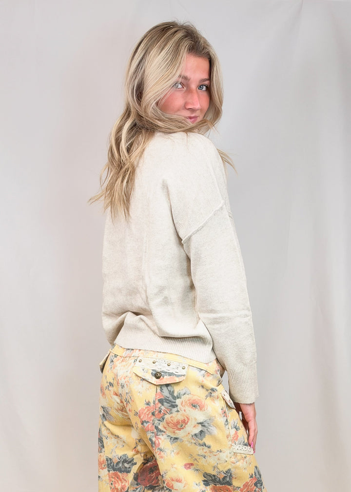 A woman stands barefoot in a studio wearing loose, baggy cream-colored pants with a colorful floral and butterfly print, paired with a light beige sweater.
