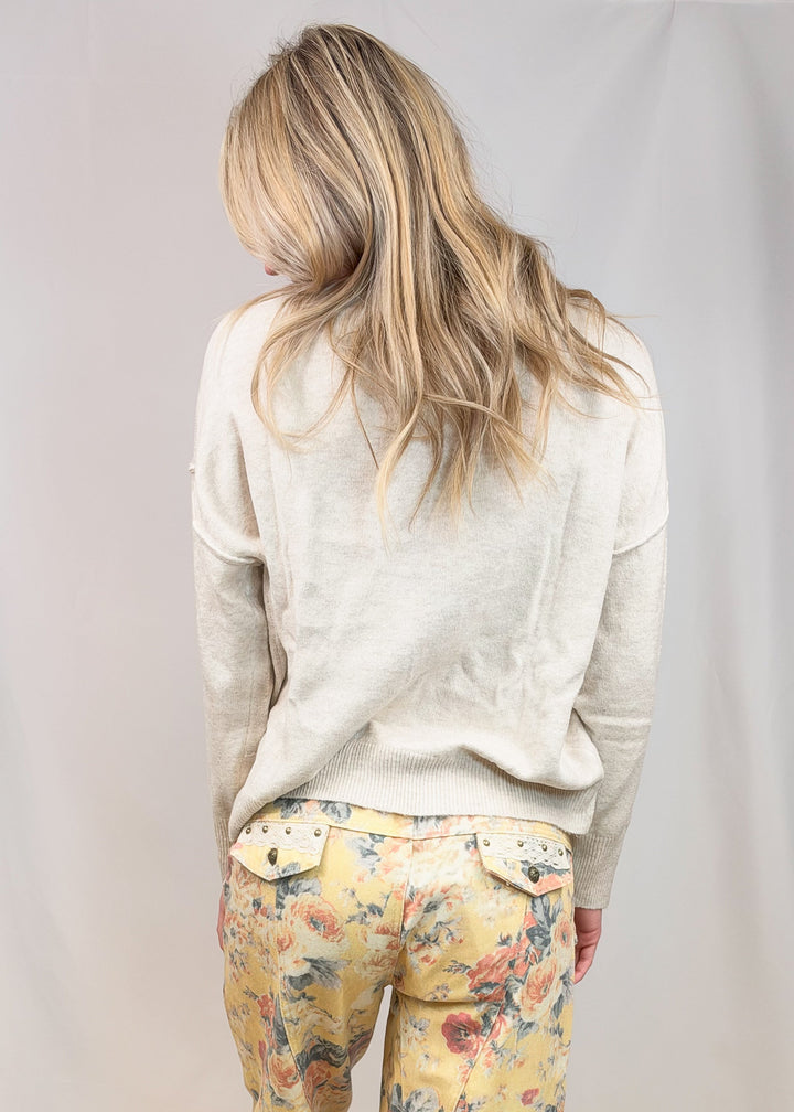 A woman viewed from behind wearing a light beige long-sleeve top and yellow floral print bohemian-style pants with ruffled hems, standing barefoot against a neutral studio backdrop.