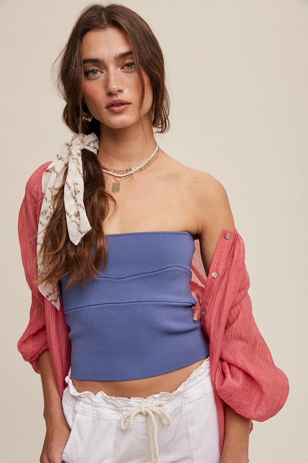 A woman modeling a strapless dusty blue crop top, styled with a pink button-down shirt draped off her shoulders and white drawstring pants, set against a solid beige background.