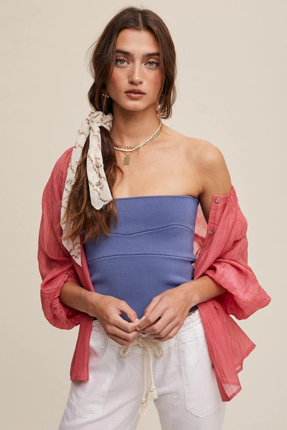A woman modeling a strapless dusty blue top paired with an open coral pink button-up shirt and white drawstring pants, against a solid beige background.