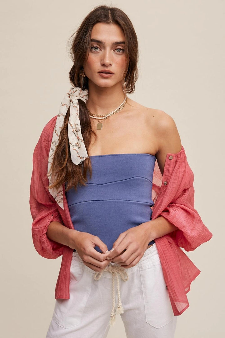A woman modeling a strapless dusty blue top paired with an open coral pink button-up shirt and white drawstring pants, against a solid beige background.