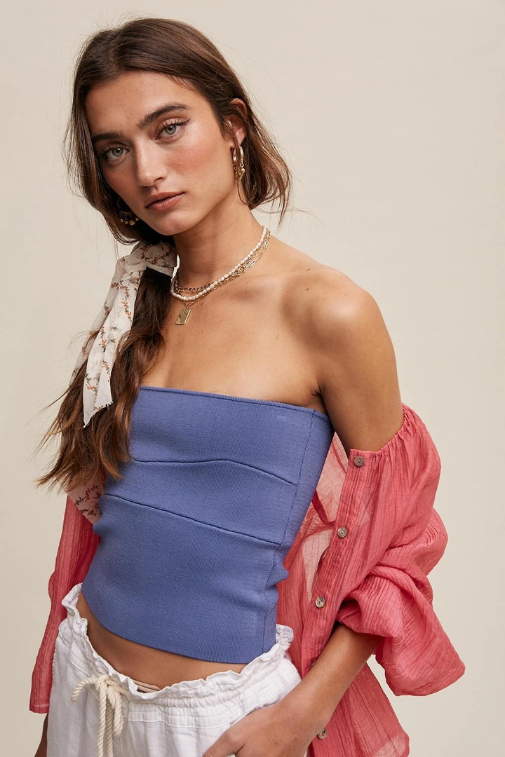 A woman modeling a blue strapless tube top, styled with a pink button-up shirt and white drawstring shorts against a solid beige background.