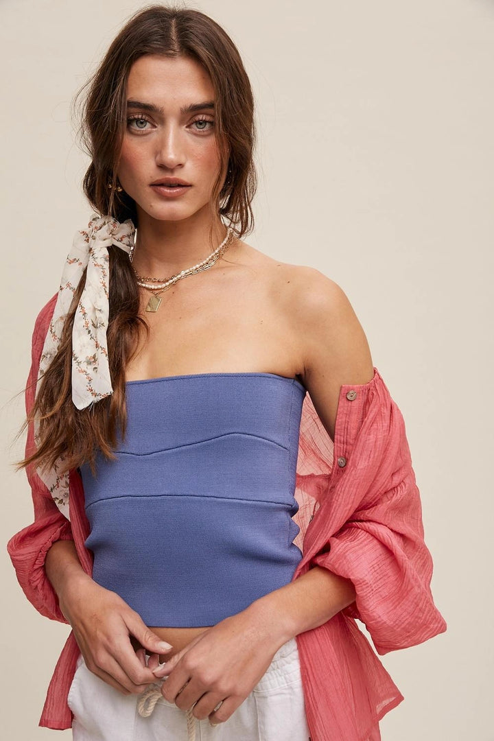 A woman modeling a strapless blue tube top with a pink button-up shirt draped off her shoulders and a cream floral neck scarf, set against a solid beige background.