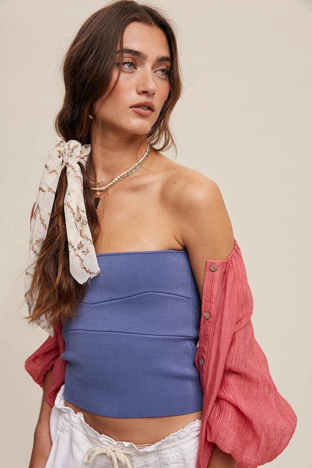 A woman modeling a blue strapless tube top with a pink button-down shirt draped off her shoulders and a cream patterned hair tie, set against a solid beige background.