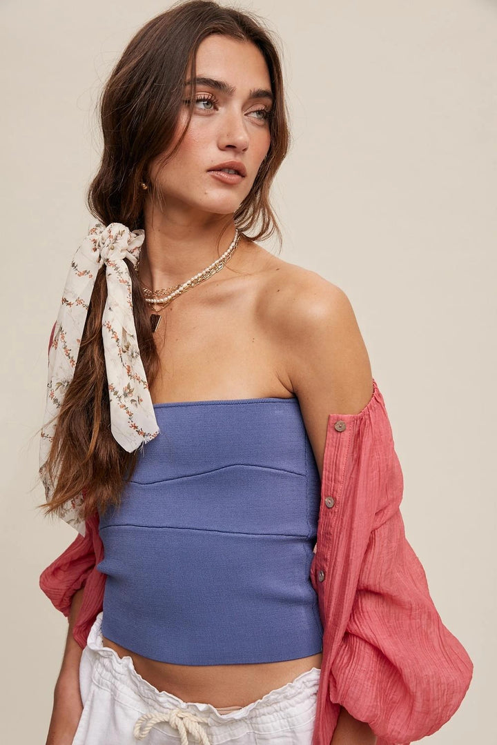 A woman modeling a blue strapless tube top with a pink button-down shirt draped off her shoulders and a cream patterned hair tie, set against a solid beige background.