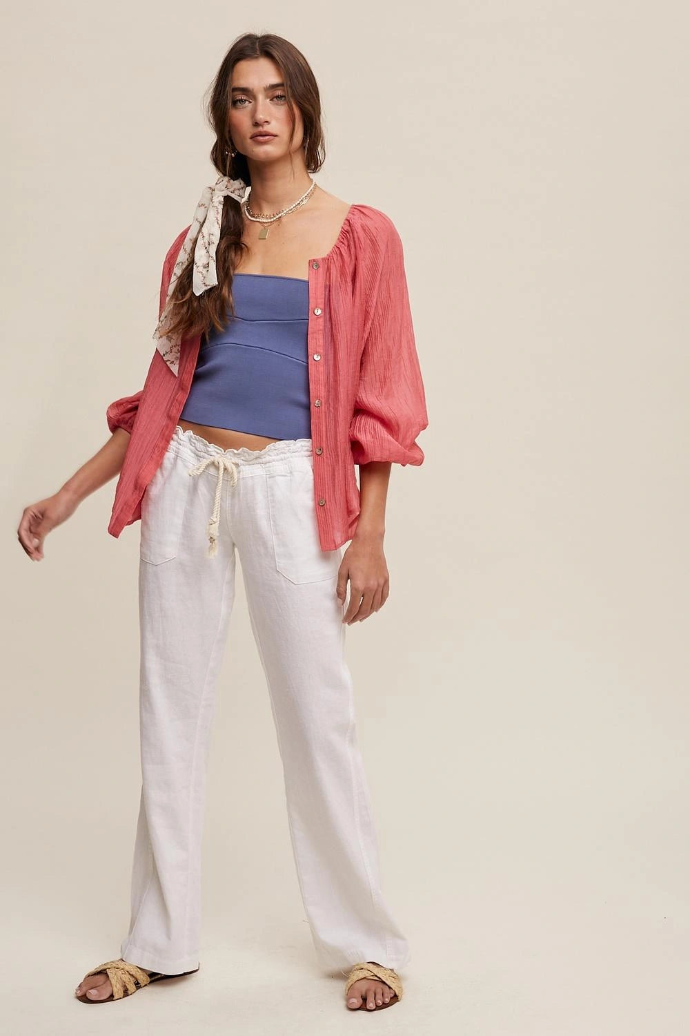 A model wearing a pink button-up shirt with puff sleeves, a blue strapless top, and white wide-leg pants, standing against a solid beige background.