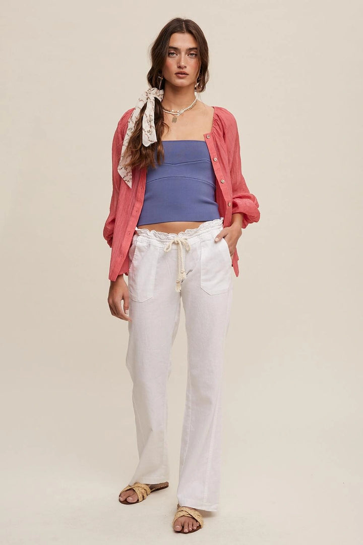 A model stands against a plain beige studio background wearing a pink button-up shirt draped open over a blue tube top, white drawstring wide-leg pants, and tan sandals. She is also wearing layered necklaces and a patterned scarf tied under her chin.