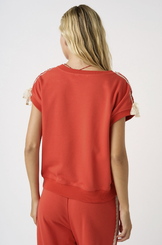 A woman shown from the back wearing a rust-red short-sleeve top with cream lace details on the sleeves and matching rust-red pants, against a plain light gray studio background.