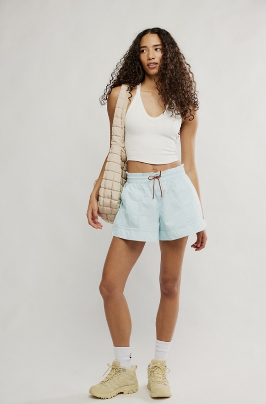 A woman modeling a light blue denim drawstring shorts, a white ribbed tank top, a beige quilted shoulder bag, white socks, and beige sneakers against a plain light gray studio background.