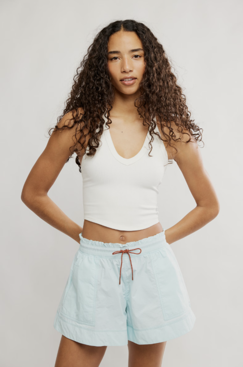 A woman modeling a white ribbed tank top and light blue drawstring shorts against a plain light gray studio background.