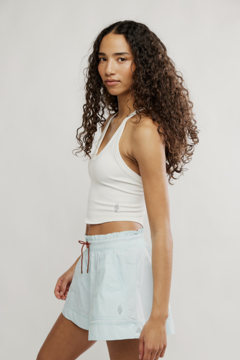 A woman with long, curly brown hair is shown in profile wearing a white ribbed crop tank top and light blue denim shorts against a plain light gray studio background.